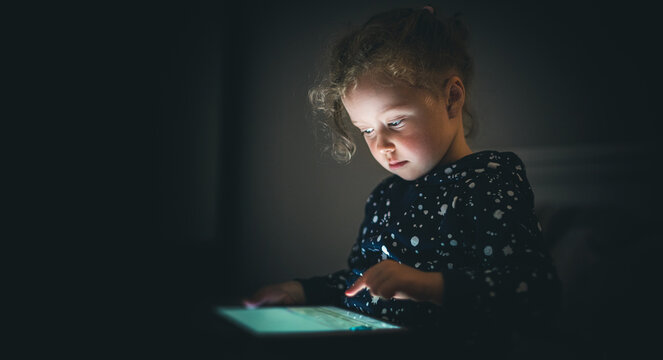 Child girl with tablet in a dark room. The baby's face is illuminated by a gadget.