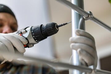 A worker in gloves repairs a metal railing of a staircase using a screwdriver and a drill