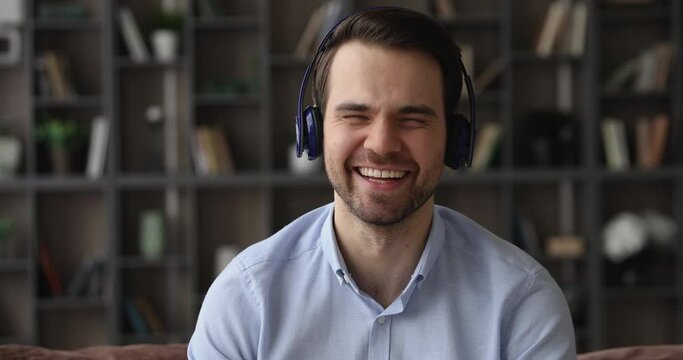 Head shot smiling handsome young man in wireless headphones looking at camera, involved in pleasant distant conversation, passing remote job interview with hr manager or communicating by video call.