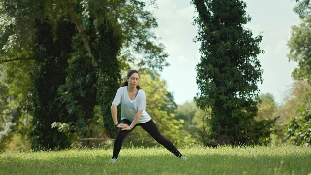 A young caucasian woman in sportswear performs warm-up squats in the summer sunny park. Slow motion.