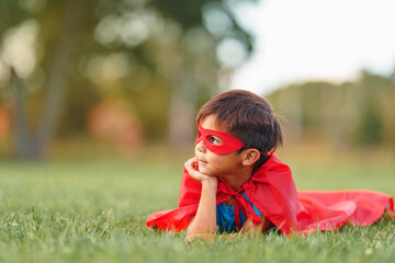 small Latin American boy in a superhero costume with a red cape is lying on the lawn in the fresh air. The child fantasizes and pretends to be a superhero. A place for text or advertising.