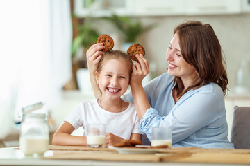 Obraz na płótnie Canvas happy loving family, mom and daughter, playing sitting at table and having breakfast at home in morning. woman and girl eat oatmeal cookies and drink cow's milk, and have nice time together in kitchen