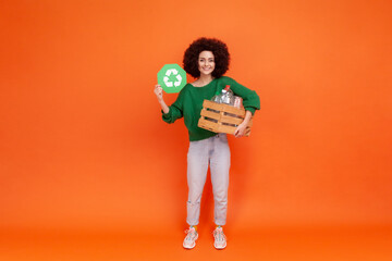 Full length portrait of woman with Afro hairstyle wearing green casual style sweater holding box with empty plastic bottler and showing green sign. Indoor studio shot isolated on orange background.