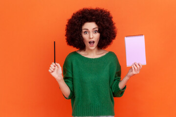 Surprised woman with Afro hairstyle wearing green casual style sweater standing with open mouth, holding paper notebook and pencil. Indoor studio shot isolated on orange background.