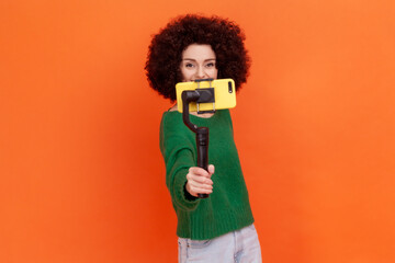 Young adult woman blogger with Afro hairstyle wearing green casual style sweater holding out steadicam with phone, making video or has livestream. Indoor studio shot isolated on orange background.
