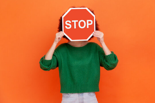 Unknown woman with wearing green casual style sweater hiding her face behind red stop traffic symbol, warning, avoids forbidden actions. Indoor studio shot isolated on orange background.