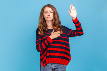 I promise. Portrait of beautiful woman wearing striped sweater, making swearing gesture and holding arm on chest, taking oath, pledging allegiance. Indoor studio shot isolated on blue background.