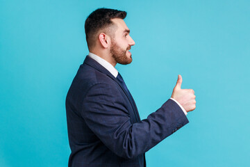 Well done, good job! Profile portrait of smiling happy bearded man wearing dark official style suit, looking ahead, showing thumb up. Indoor studio shot isolated on blue background.