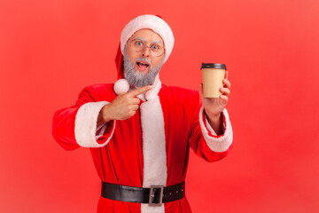 Fototapeta na wymiar Elderly man with gray beard wearing santa claus costume pointing at coffee, drink with caffeine, energy boost in morning, looking at camera. Indoor studio shot isolated on red background.