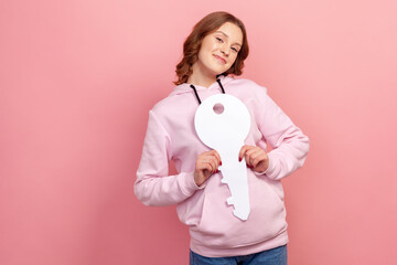 Portrait of happy curly haired teenage girl in hoodie holding paper key as symbol of own apartment, dreams comes true, state housing program. Indoor studio shot isolated on pink background