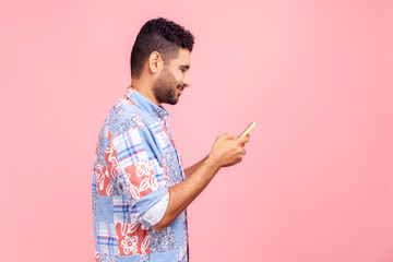 Side view of handsome dark haired man with beard with wearing casual style shirt using online service on mobile phone and expressing pleasant emotions. Indoor studio shot isolated on pink background.