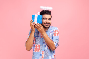 Smiling positive man with nimb over head looking at camera with happy expression, holding wrapped present box in hands, looking at camera. Indoor studio shot isolated on pink background.