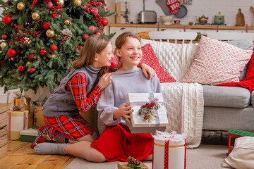 Obraz na płótnie Canvas Two joyful sisters are sitting at home near christmas tree and looking at boxes with gifts