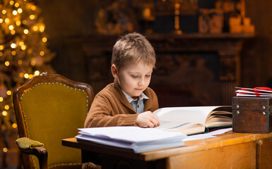 Boy reads a magic book while sitting at the table. Home interior with Christmas tree and fireplace. Traditional Christmas concept.