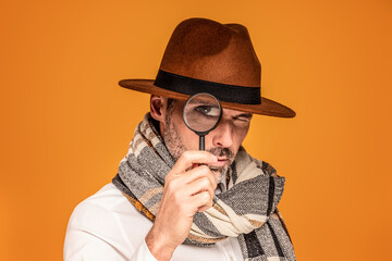 Male detective with magnifying glass, wearing fashionable scarf and brown hat, posing on yellow...