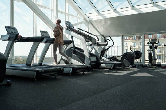 Horizontal long shot of modern Muslim woman wearing hijab exercising in gym running on treadmill, copy space