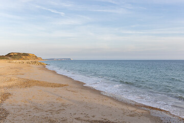 A scenic beautiful view of a sandy beach with blue sea, rocky groyne, majestic hills and golden hours colors under a majestic blue sky and some white clouds