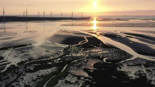 Cinematic aerial shot at Gaomei wetland preservation area capturing the distinctive grass marsh coastal wetland pattern and texture at sunset, renewable energy wind turbines spinning at the horizon.