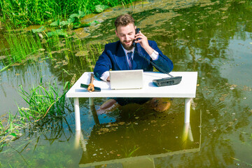 A man in a jacket sits waist-deep in the river at a table in front of a laptop and holds a phone near his ear