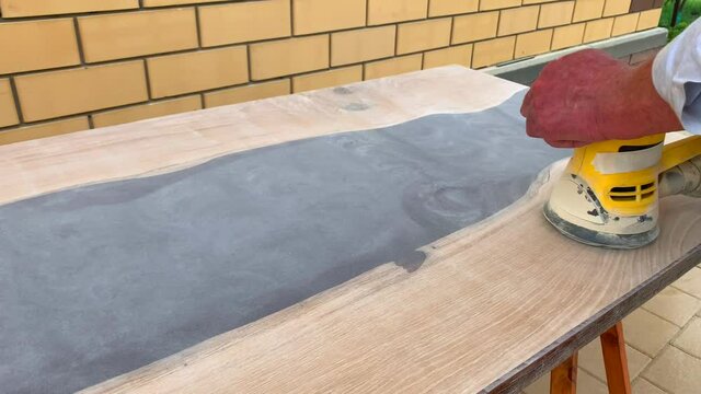 Grind table with polishing machine close-up. man works epoxy table. Work hands wooden table. Close-up of man's hands of builder. DIY work on street. Polish in circular motion in shirt. River table.