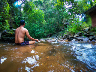 man meditating in natural waterfall with green forests from low angle