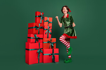 Photo of impressed lucky young woman dressed elf costume standing boxes pile rising fists smiling...