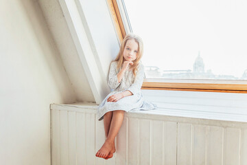 Little cute sweet smiling girl in white dress sitting on window sill in bright light living room at home indoors. Childhood schoolchildren youth relax concept.