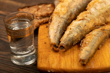 A small fried fish on a wooden board, pieces of bread and a glass of vodka, close-up, selective focus.