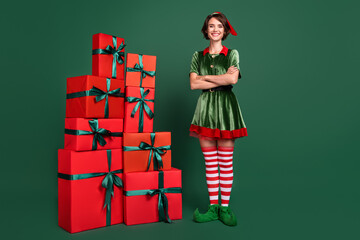 Photo of pretty confident young woman dressed elf costume standing gift boxes pile arms crossed...