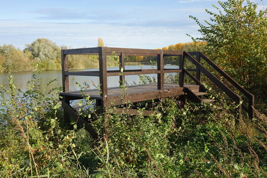 Wooden outlook platform amidst bushes at the waterfront of Blaue Adria recreational area and natural habitat on an autumn day, Altrip Germany
