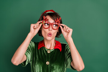 Photo of pretty shocked young woman dressed elf costume deer glasses looking empty space isolated green color background