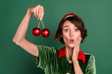 Photo of amazed shocked lady hold two baubles open mouth wear elf costume hat isolated green color...