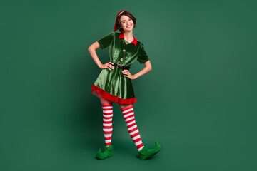Photo of funny positive cheerful helper lady hands hips wear elf costume hat stockings isolated green color background - obrazy, fototapety, plakaty