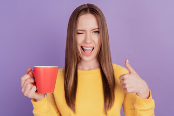 Portrait of reliable lady hold mug drink coffee wink eye raise thumb up on purple background