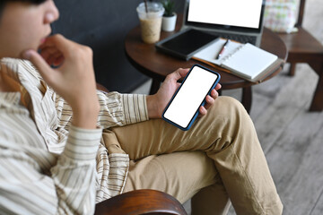 Obraz premium Closed up with young man keeping hand on chin while holding a white blank screen smartphone at the wooden chair.