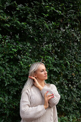 blonde woman in sweater, standing against background of wall of green leaves, hands near her face, eyes closed.