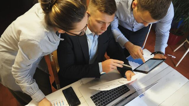 Close up to group of employees comparing sales result graph on paper with chart on tablet pc screen at office. Young colleagues examining financial reports during business meeting indoor. Slow motion