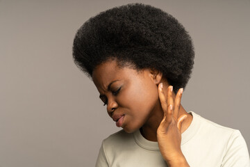 Unhealthy young african american woman frowning, touches and checking lymph nodes with finger, suffering from ear pain, isolated on studio gray background. Earache, otitis disease concept