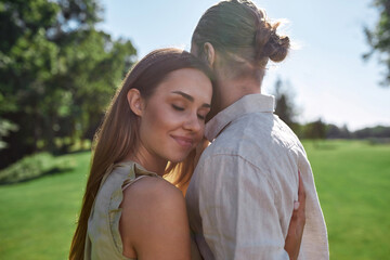 Portrait of cute young woman smiling with eyes closed, embracing her husband, posing together outdoors while standing in the park