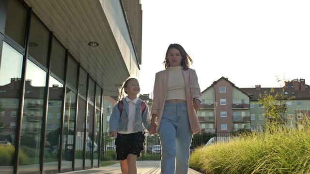 Young woman leads her daughter 6-7 years old to school by the hand. Girl with pigtails happily tells her mother something. Back to school.