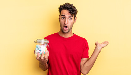 hispanic handsome man looking surprised and shocked, with jaw dropped holding an object. candies...