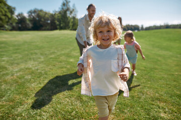 Joyful little boy smiling while playing catch together with his parents and sister in green park on a summer day. Happy family enjoying leisure activity