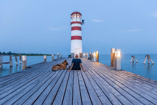 Man with DogWatching Sunrise at Wooden Pier with Lighthouse .  Togetherness and Friendship in Travel