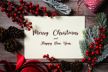 Merry Christmas and happy New Year card. Natural festive holiday composition. Branches, red berries, twine, pine cones, ribbon. Text on white card on dark wooden background. Holiday greeting.