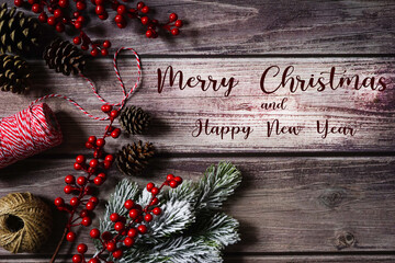 Merry Christmas and happy New Year card. Natural festive holiday composition. Branches, red berries, twine, pine cones. Text on dark wooden background. Holiday greeting.