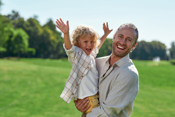 Loving young dad smiling at camera, holding his child while playing with him on grass field on a summer day. Little boy raised his arms, looking cheerful