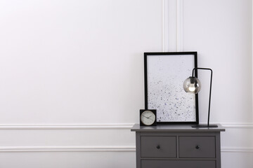 Grey wooden chest of drawers with lamp, clock and picture near white wall in room, space for text. Interior design