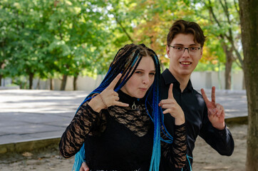 A young couple showing victory gestures with their hands against the yellow-green trees. Man and Woman with long blue pigtails. Young adults grimace. Love, relationships. Autumn city park.
