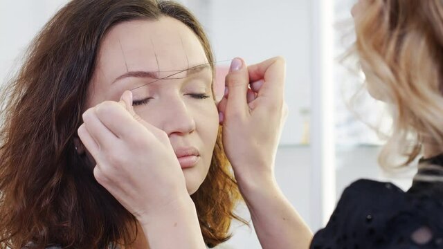 Cosmetologist forms the eyebrow line with a black thread on a female face. Makeup artist draws the shape of a woman's eyebrows in a beauty salon.
