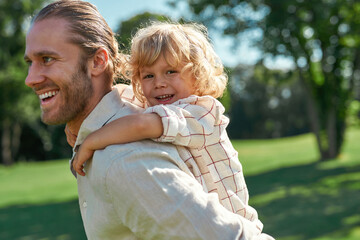 Portrait of cheerful little boy playing in the park with his father giving son piggyback ride on...